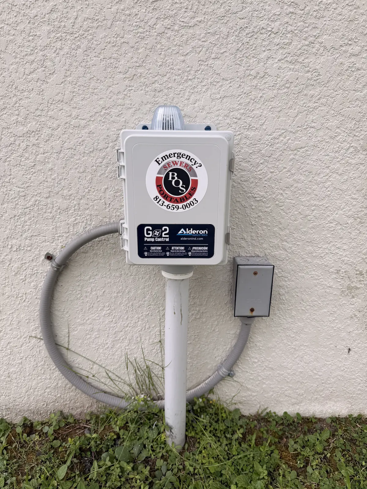 G2 lift station alarm panel mounted on a house wall, with a Bingham On-Site Emergency 813-659-0003 sticker