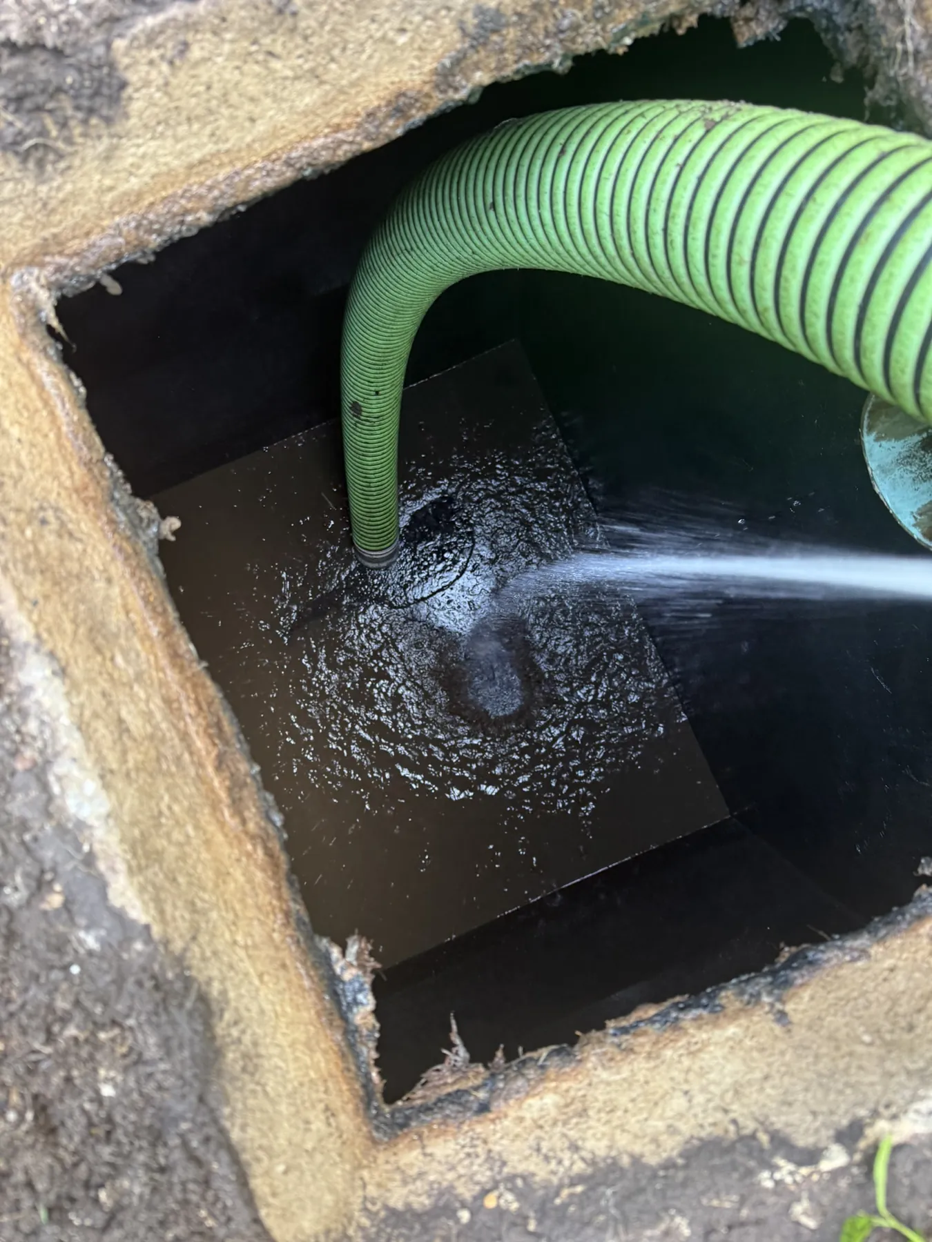 Open concrete septic tank being pumped with a green suction hose while a water jet breaks up solids